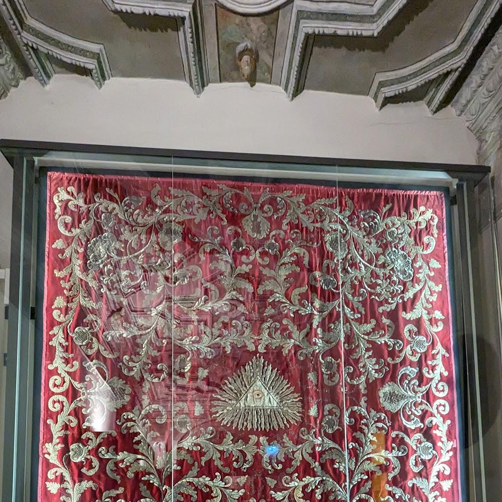 A red fabric tapestry with gold embroidery in a glass case.  The tapestry has a gold eye with rays around it.  The fabric is framed in a dark wood frame.